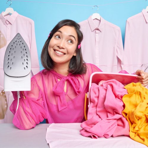 Dreamy smiling young brunette Asian woman concentrated above going to do ironing at home holds steam electric iron thinks about plans after finishing housework. Laundering and domestic work concept Dreamy smiling young brunette Asian woman concentrated above going to do ironing at home holds steam electric iron thinks about plans after finishing housework. Laundering and domestic work concept